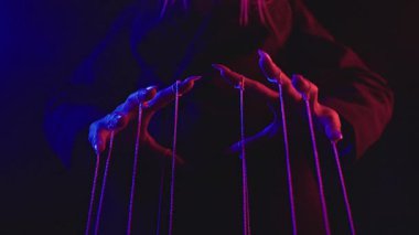 Uzun tırnaklı gizemli bir figür dramatik neon ışıkları altında ipleri manipüle ederek ürkütücü bir atmosfer yaratıyor. Yavaş çekim