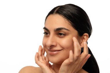 Gülümseyen bir kadın nazikçe yüzüne dokunur, rahatlama ve cilt bakımını ön plana çıkarır. İfade güven ve pozitiflik taşır..