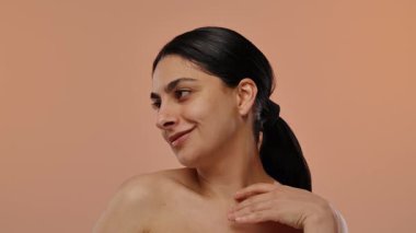 Siyah saçlı, kendine güvenen bir kadın şeftali rengi bir arka planda hoş bir gülümsemeyle poz verir. Yavaş çekim