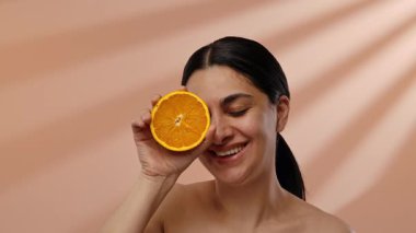 Bir kadın gözünün önünde bir dilim portakalı tutarken, sıcak bir bej arka plana yaslanarak geniş bir şekilde gülümsüyor. Yavaş çekim