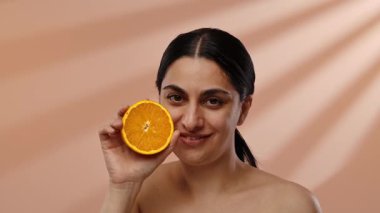 Bir kadın elinde portakal dilimi tutarken gülümsüyor, güneş ışığına karşı, şeftali rengi bir arka plan, sıcak ve sağlıklı bir hava yaratıyor. Yavaş çekim