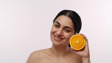 Koyu saçlı bir kadın yüzünün yanında turuncu bir şey tutuyor ve taze narenciye sergiliyor. Arka plan düz beyaz. Yavaş çekim