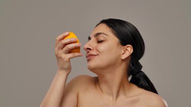 Parlak bir gülüşü olan bir kadın, minimalist bir ortamda yarı kesilmiş turuncu bir güzellik ve tazelik sergiliyor. Yavaş çekim