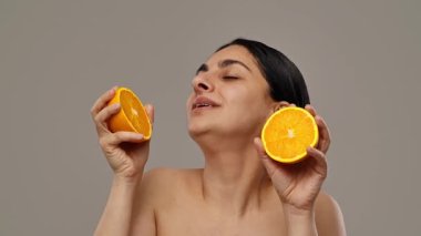 Neşeli bir kadın taze portakal yarımları tutuyor, tarafsız bir arkaplanda mutluluk ve sağlık yayıyor. Yavaş çekim