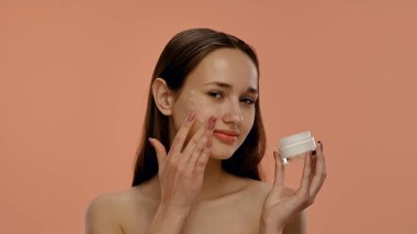 Bir kadın elinde bir kavanoz krema tutarken gülümser, şeftali arka planına yanaklarına sürerek.