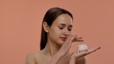 Bir kadın elinde bir kavanoz krema tutarken gülümseyip şeftali arka planına yanaklarına sürüyor. Yavaş çekim