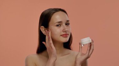 Bir kadın elinde bir kavanoz krema tutarken gülümseyip şeftali arka planına yanaklarına sürüyor. Yavaş çekim