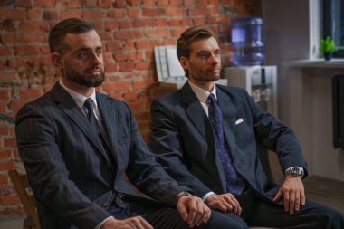 Resmi takım elbiseli iki işadamı, duvarları tuğlalı modern bir ofiste oturmuş, bir tartışma içerisindeler..