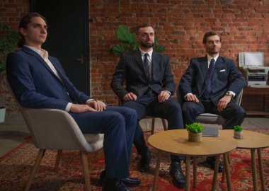 Resmi iş kıyafeti giymiş üç adam modern bir ofis ortamında oturmuş mesleki meseleleri tartışıyorlar..