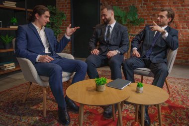 Resmi iş kıyafeti giymiş üç adam modern bir ofis ortamında oturmuş mesleki meseleleri tartışıyorlar..