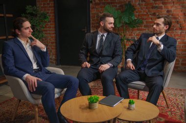 Resmi iş kıyafeti giymiş üç adam modern bir ofis ortamında oturmuş mesleki meseleleri tartışıyorlar..