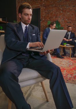 Takım elbiseli bir işadamı modern ofis ortamında iş arkadaşlarıyla iş hakkında konuşan bir dizüstü bilgisayar kullanıyor..