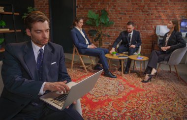 Takım elbiseli bir işadamı modern ofis ortamında iş arkadaşlarıyla iş hakkında konuşan bir dizüstü bilgisayar kullanıyor..