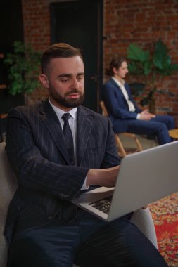 Takım elbiseli bir işadamı modern ofis ortamında iş arkadaşlarıyla iş hakkında konuşan bir dizüstü bilgisayar kullanıyor..