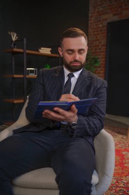 İnce çizgili takım elbiseli bir adam, açık tuğla duvarlı rahat bir ofiste otururken deftere yazıyor..