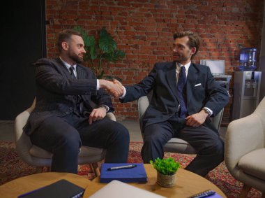 Takım elbiseli iki işadamı modern bir ofiste tuğla duvar ve saksılarla el sıkışıyor ve profesyonel işbirliğini vurguluyorlar..