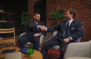 Takım elbiseli iki işadamı modern bir ofiste tuğla duvar ve saksılarla el sıkışıyor ve profesyonel işbirliğini vurguluyorlar..