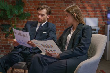 Resmi giyinmiş iki iş adamı finansal tabloları analiz ediyor ve modern ofis ortamında strateji tartışıyorlar..