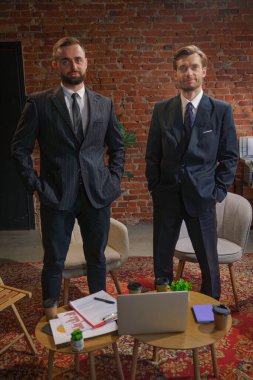 Takım elbiseli iki kendine güvenen işadamı, modern ofis ortamında ellerinde belgeler ve kahveyle bir masanın başında duruyorlar..