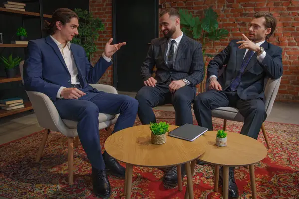 Resmi iş kıyafeti giymiş üç adam modern bir ofis ortamında oturmuş mesleki meseleleri tartışıyorlar..