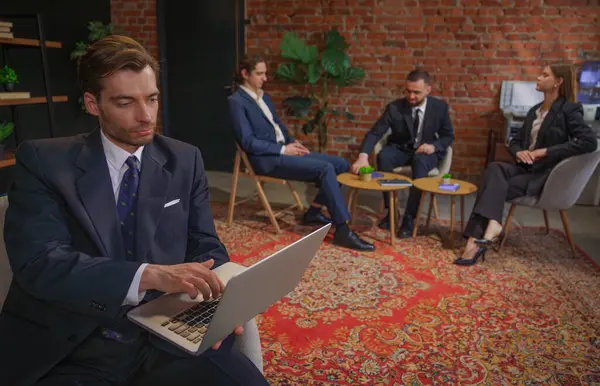 Takım elbiseli bir işadamı modern ofis ortamında iş arkadaşlarıyla iş hakkında konuşan bir dizüstü bilgisayar kullanıyor..