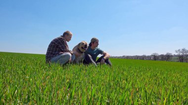 Doğa ve manzaralı bir köy manzarası ile çevrili çimenli bir tarlada köpeğiyle güneşli bir günün tadını çıkaran iki insan..