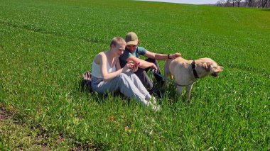 İki arkadaş yemyeşil bir alanda mutlu bir köpekle oturuyorlar. Açık mavi gökyüzünün altında birbirlerine eşlik ederek eğleniyorlar ve telefonlarına bakıyorlar.