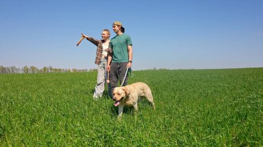 İki arkadaş yemyeşil bir alanda mutlu bir köpekle yürüyorlar. Açık mavi gökyüzü altında birbirlerine eşlik etmekten hoşlanırlar..