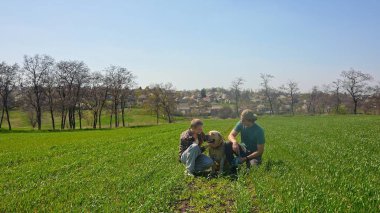 İki arkadaş yemyeşil bir alanda mutlu bir köpekle oturuyorlar. Berrak, mavi gökyüzünün altında birbirlerine eşlik ederek eğleniyorlar..