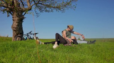 Bir çift, yakınlarda bisikletleri olan sakin yeşil bir tarlada bir ağacın altında oturuyor. Güneşli bir gün huzurlu bir sahne yaratır..