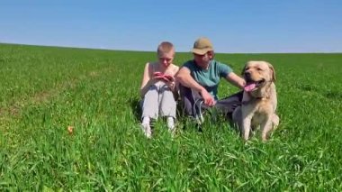 İki arkadaş yemyeşil bir alanda mutlu bir köpekle oturuyorlar. Açık mavi gökyüzü altında birbirleriyle eğleniyorlar ve telefonlarıyla oyun oynuyorlar.
