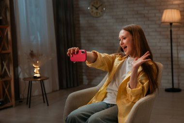 Günlük yaşam tarzı konsepti. Genç kız akşam vaktini evde geçiriyor, kız oturma odasında oturuyor ve akıllı telefondan selfie çekiyor, komik suratlar yapıyor ve gülümsüyor..