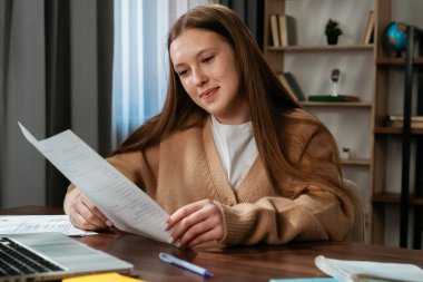 Günlük yaşam tarzı konsepti. Genç kız evde ders çalışıyor, kız dizüstü bilgisayar ve kitaplarla masaya oturuyor, defterde kalem ve kalemle ödev çiziyor, ekrana bakıyor..
