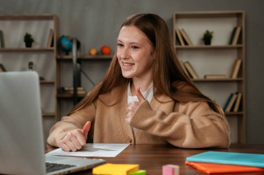 Gülümseyen genç bir öğrenci, olumlu ve ilgi çekici bir deneyimi sembolize eden bir video görüşmesi sırasında başparmağını kaldırır..