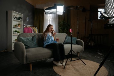 Sahne arkasında. Günlük yaşam tarzı konsepti. Genç kız evde vakit geçiriyor, kız blogcu kanepeye oturuyor akıllı telefondaki videoyu kaydediyor, kameraya konuşuyor kozmetik tozunu gösteriyor, yüzüne fırça sürüyor..