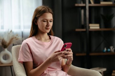 Pembe gömlekli genç bir kadın rahat bir şekilde sandalyeye oturur, bir mesaj yazar ve video izler..