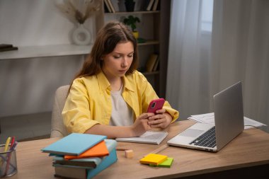 Genç bir kadın, kitaplarla çevrili bir akıllı telefon ve dizüstü bilgisayar kullanarak bir masada derslerine odaklanıyor..