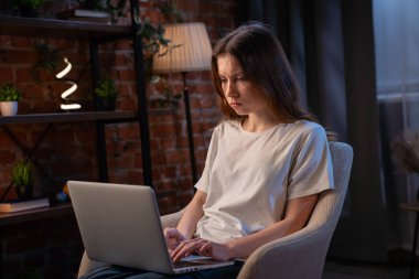 Genç bir kadın sıcak ışıklandırmasıyla rahat bir evde otururken dikkatle dizüstü bilgisayarına odaklanıyor..