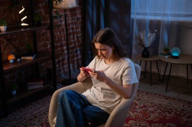 Genç bir kadın, modern dekorlu sıcak, aydınlık bir odada otururken telefonuna sinirlenmiş bakar..