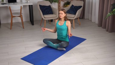Genç bir kadın, içeride mavi bir minder üzerinde yoga egzersizleri yapıyor, esneklik ve gevşemeye odaklanıyor..