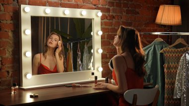 Kırmızı elbiseli bir kadın gösteriş için oturur, moda ve ışıkla çevriliyken makyaj yapar..