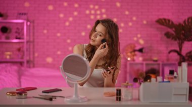 Bir kadın elinde toz fırçasıyla masada oturuyor, sıcak, pembe bir odada kozmetik aletlerle çevrili..