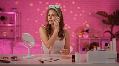 Genç bir kadın, parlak, pembe temalı bir odada makyaj aletleri ile çevrili cilt ürünleri uyguluyor..