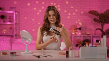 Masada oturan bir kadın elinde bir göz falı paleti, sıcak bir şekilde aydınlatılmış pembe bir odada kozmetik aletlerle çevrili..