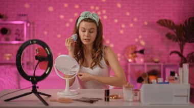 Genç bir kadın şık bir odada makyaj yapar. Güzellik aletleri ve kozmetiklerle çevrili bir ayna ve profesyonel ışıklandırma kullanıyor..