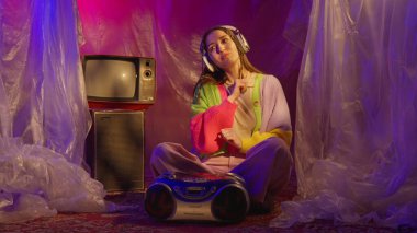 Genç bir kadın, renkli kıyafetler ve kulaklıklar takmış, müziğin tadını çıkaran, neon ışıklı, retro temalı bir odada oturuyor..