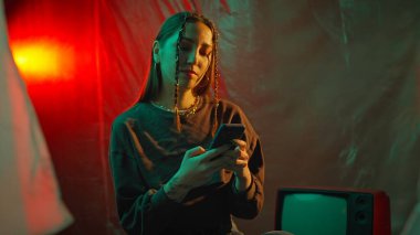 Bir kadın eski bir televizyonlu loş, modası geçmiş bir odada otururken akıllı telefonunu kullanmaya odaklanır..