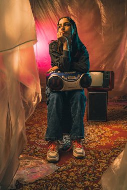 Renkli perdelerle çevrili klasik elektronik aletlerle oturan bir kadın nostaljik bir atmosfer yaratıyor..