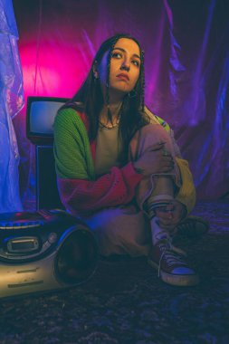 Genç bir kadın, canlı neon ışıklarıyla çevrili eski elektronik eşyaların yanında oturup nostaljik ama modern bir estetik yaratıyor..