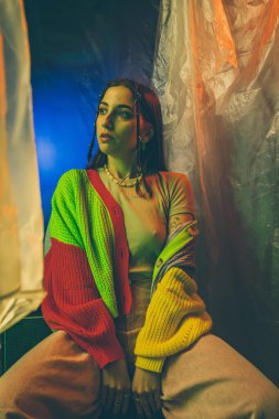 Canlı bir elbise içinde renkli bir arkaplanda poz veren bir kadın canlı neon ışıklar ve karışık dokular sergiliyor..